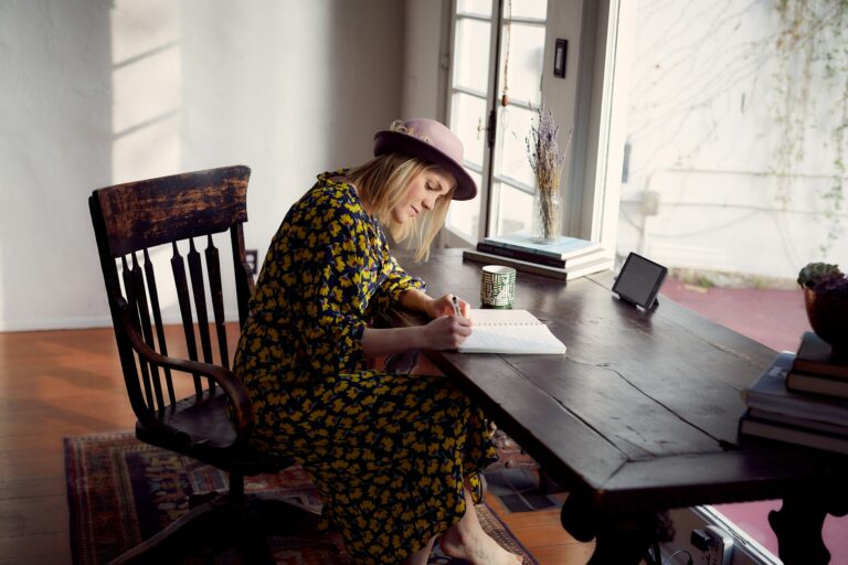 A woman wearing a hat and floral dress sits at a wooden desk in a sunlit room, hand-writing in a notebook with a pen, surrounded by books and a small tablet.