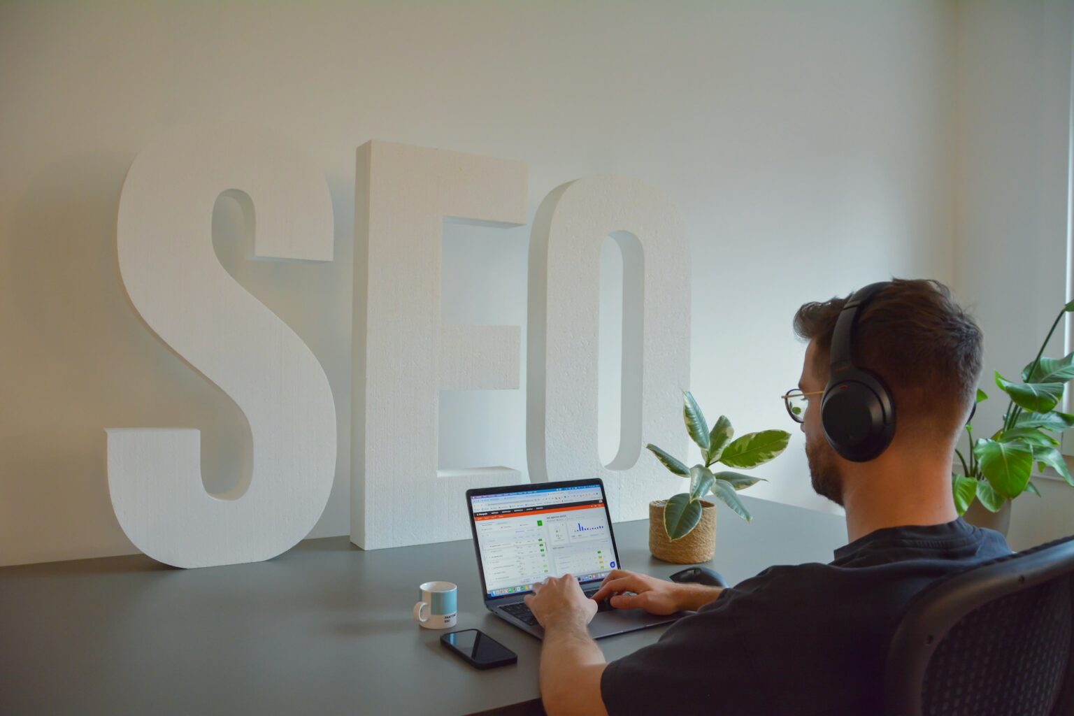 A man wearing headphones sits at a desk working on a laptop displaying analytics data, with large white 3D letters spelling 'SEO' standing prominently in the background.