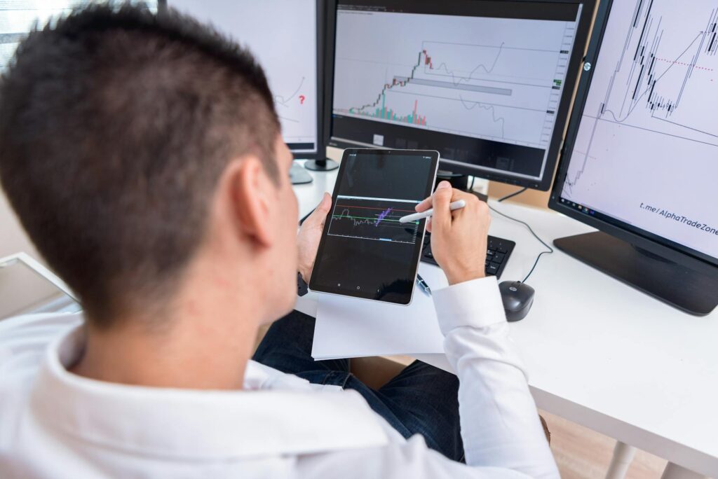 Over-the-shoulder view of a financial analyst reviewing complex data charts on a tablet and multiple computer monitors, representing financial tracking and statement analysis.