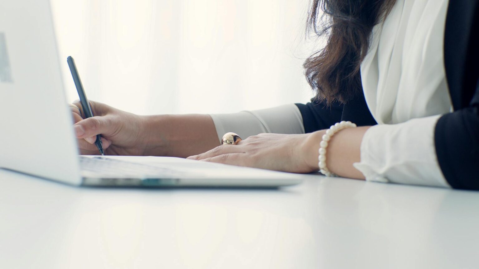 Close-up side view of a professional woman's hands writing notes with a pen next to a laptop keyboard, wearing a pearl bracelet, representing detailed budget planning or business analysis.