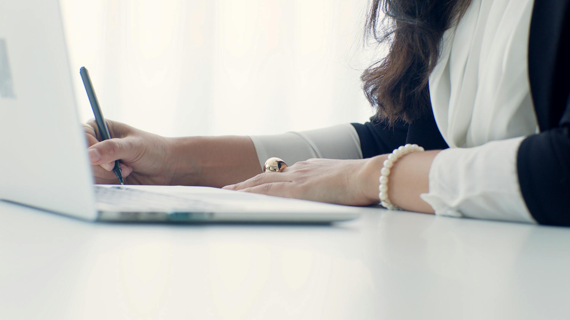 Close-up side view of a professional woman's hands writing notes with a pen next to a laptop keyboard, wearing a pearl bracelet, representing detailed budget planning or business analysis.