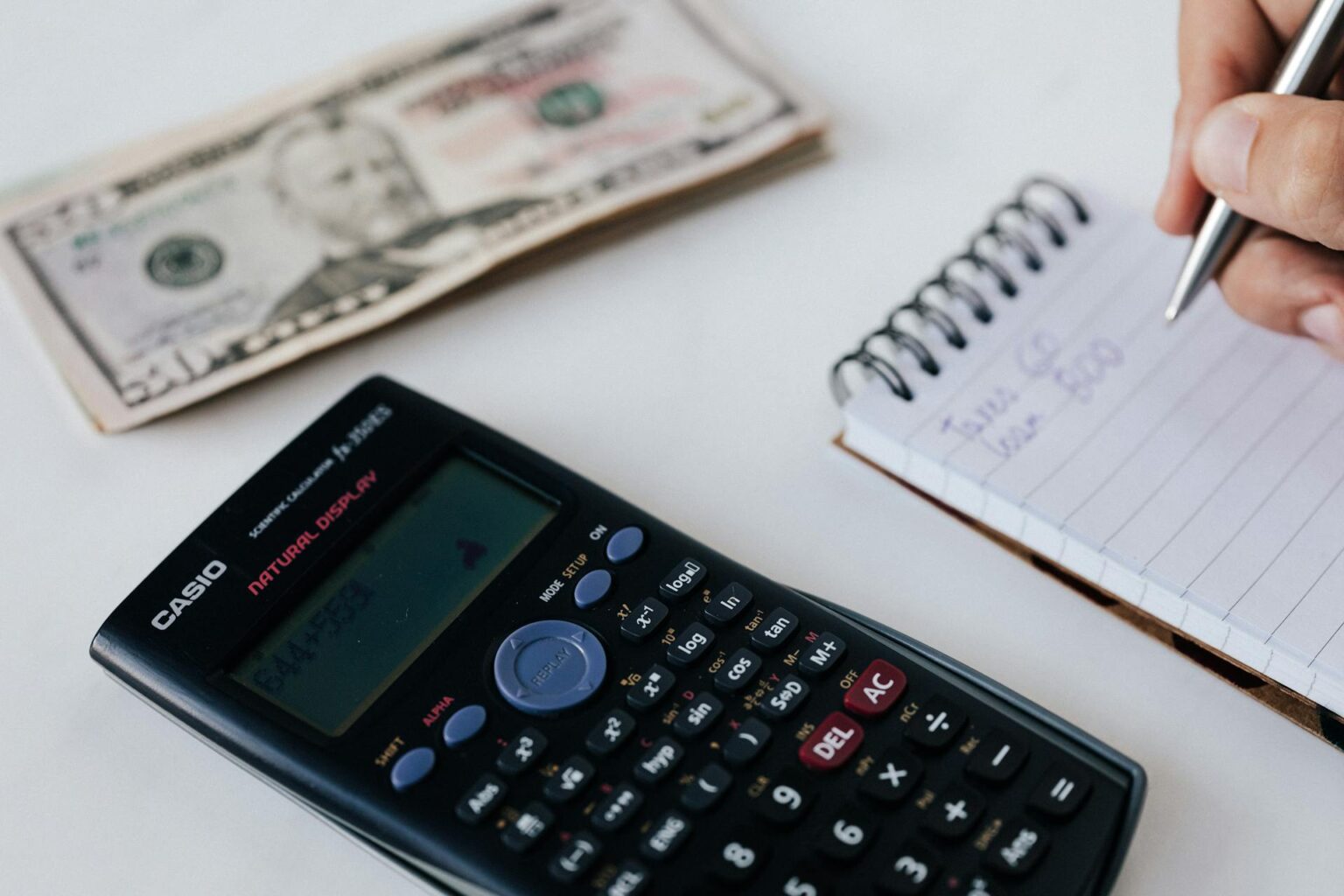 Close-up of a person's hand writing notes in a spiral notebook next to a scientific calculator and a stack of US dollar bills, representing financial planning, tax calculation, or budgeting.