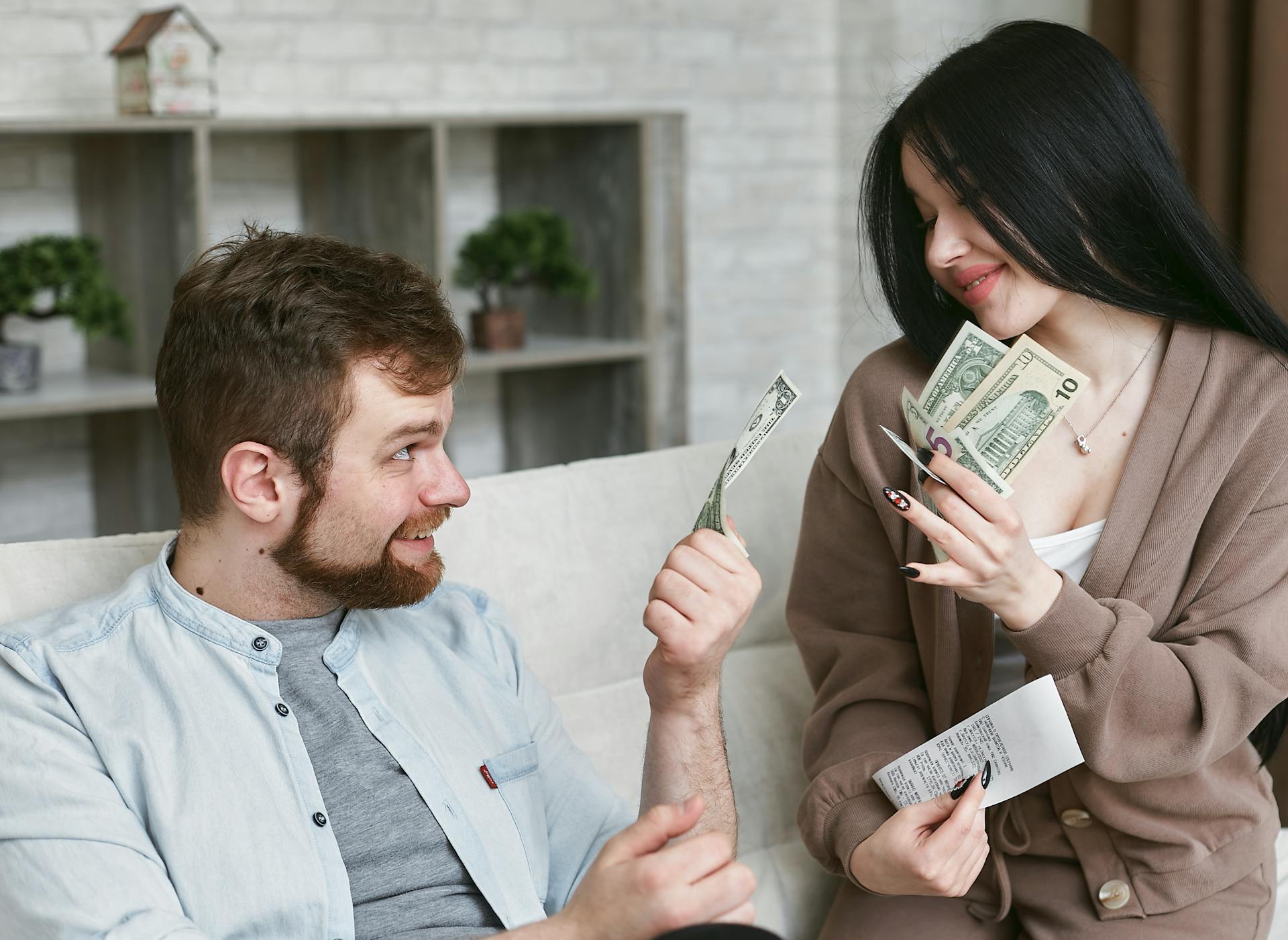 A young couple sitting on a sofa discussing their finances while holding cash and receipts, representing household budgeting and financial planning.