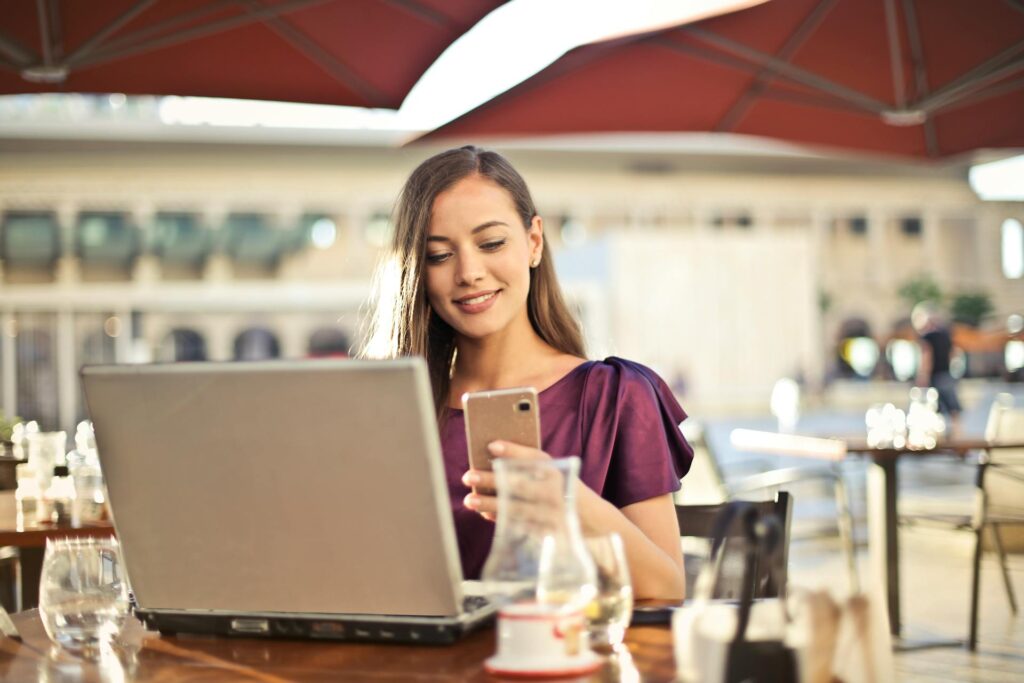Young female entrepreneur working remotely on a laptop and smartphone at an outdoor cafe, representing the flexibility of starting a small business from home.