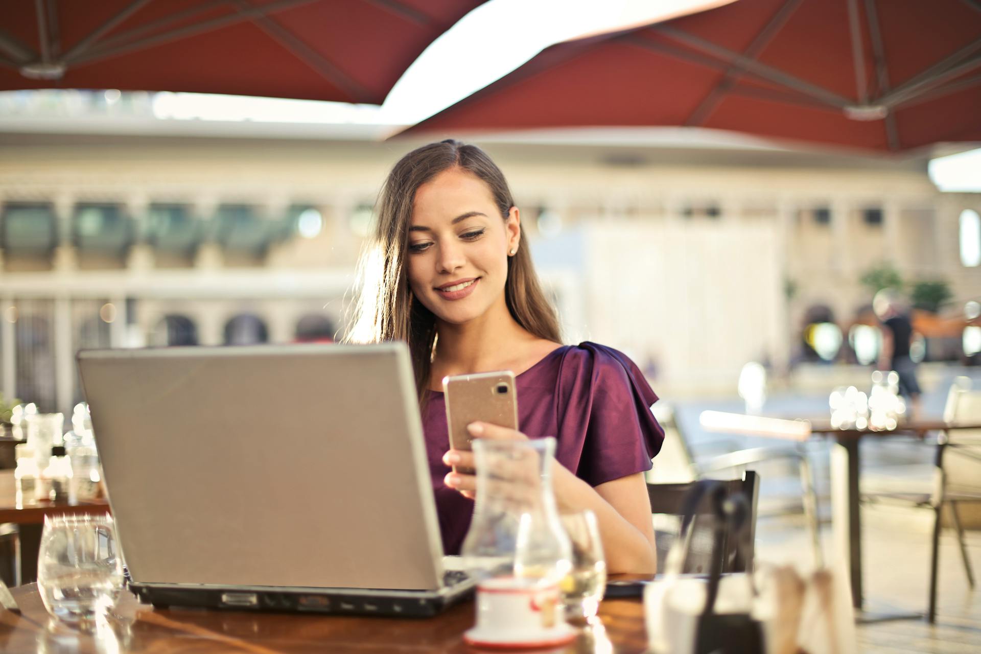 Young female entrepreneur working remotely on a laptop and smartphone at an outdoor cafe, representing the flexibility of starting a small business from home.