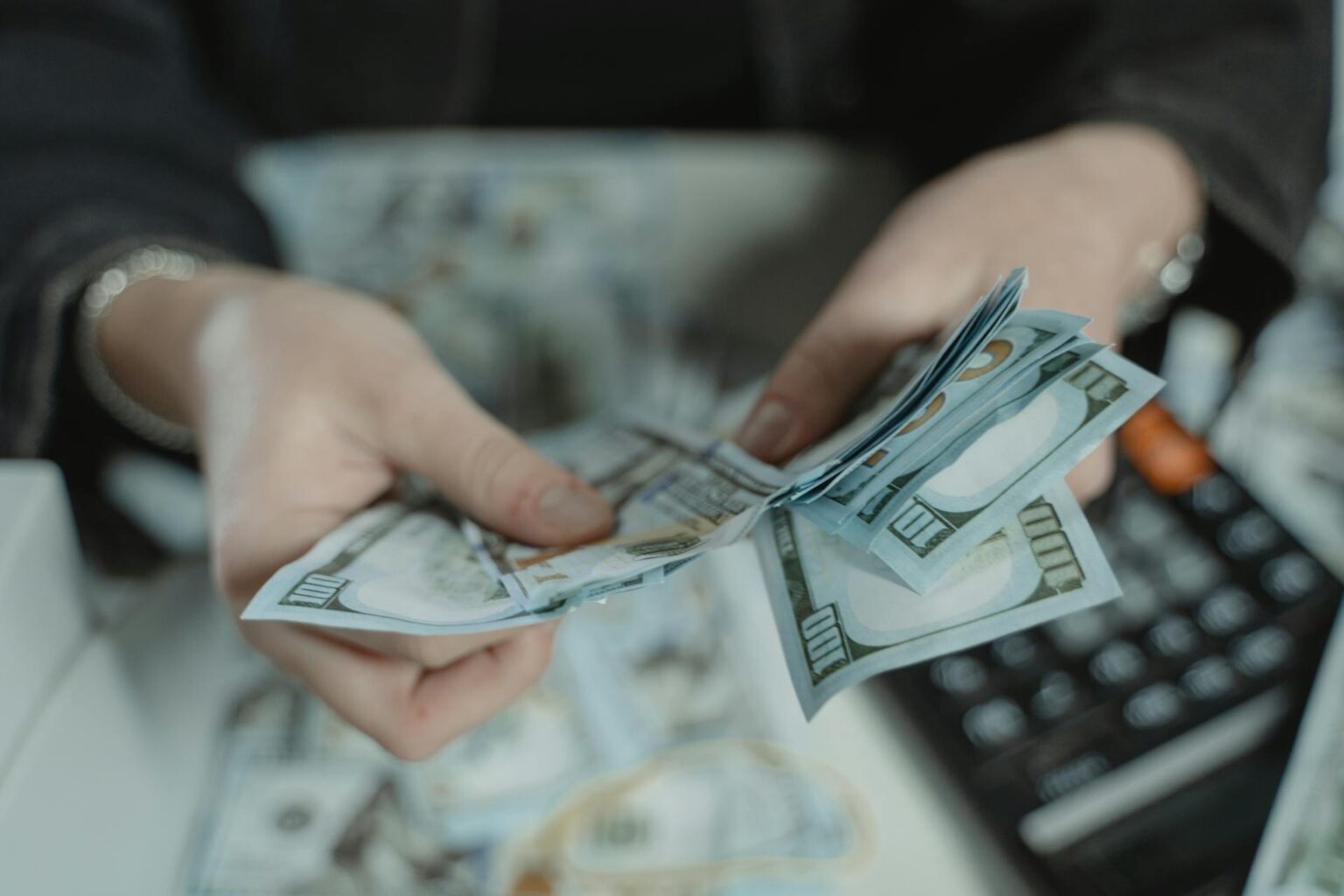 Close-up of hands counting a stack of one hundred dollar bills with a calculator in the background, representing budgeting and financial planning.
