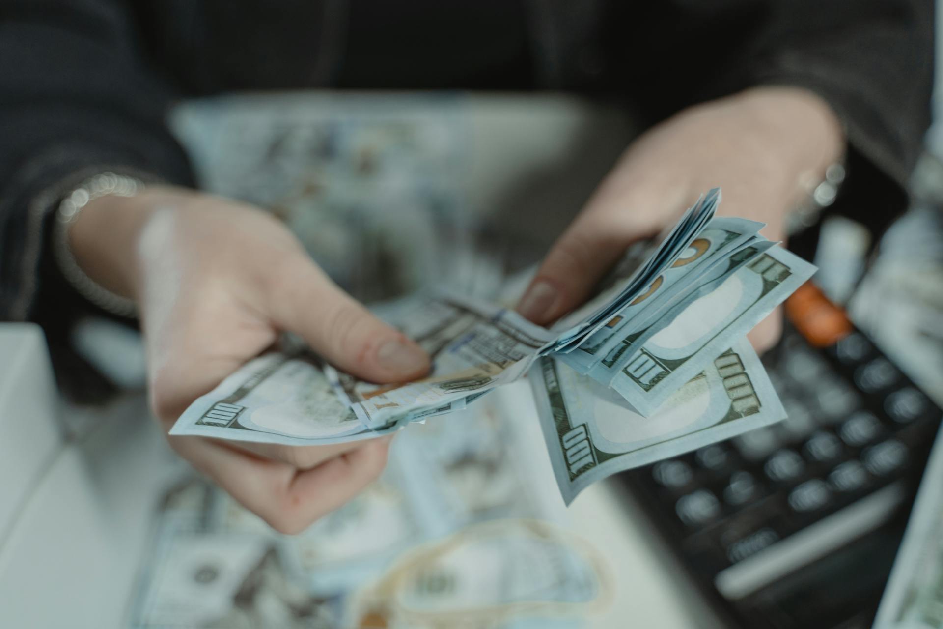 Close-up of hands counting a stack of one hundred dollar bills with a calculator in the background, representing budgeting and financial planning.